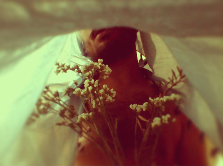 A nude man with a bedsheet over his head, is holding a bunch of small white flowers. The photo is clicked from inside the bedsheet. He is visible from the lips till the midrif.