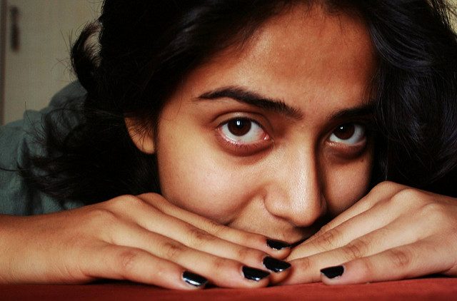 A brown-skinned woman resting her hands and her cheek on a table, looking wide-eyed straight as if peeking at something. She is wearing black nailpaint.