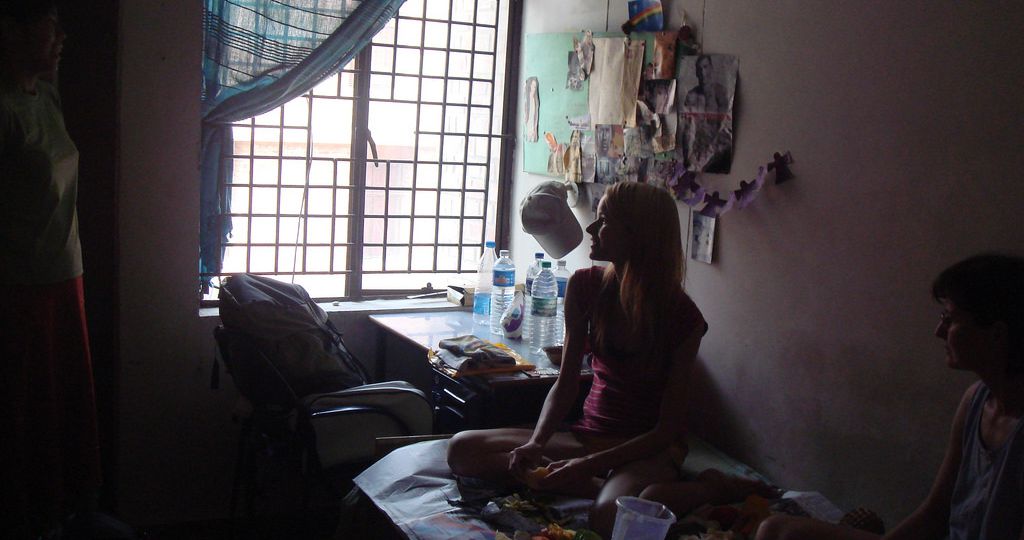 A woman sitting on a bed in her room wearing shorts and a tank top, looking out the window.