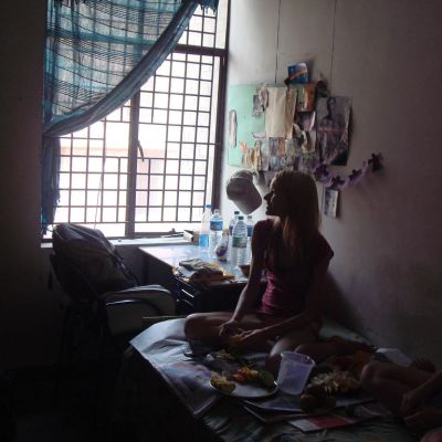 A woman sitting on a bed in her room wearing shorts and a tank top, looking out the window.