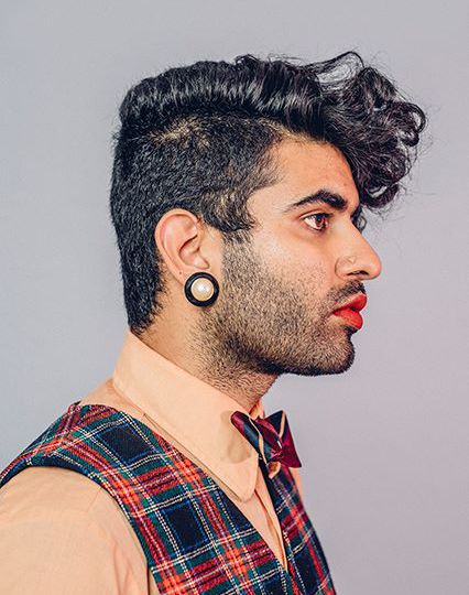 A right profile of Alok Vaid-Menon with a stub, wearing red lipstick, yellow nosering, big golden and black earrings, and hair parted by the side - with right side "manly" hair, and left - longer and curlier.