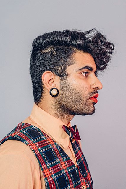A right profile of Alok Vaid-Menon with a stub, wearing red lipstick, yellow nosering, big golden and black earrings, and hair parted by the side - with right side "manly" hair, and left - longer and curlier.