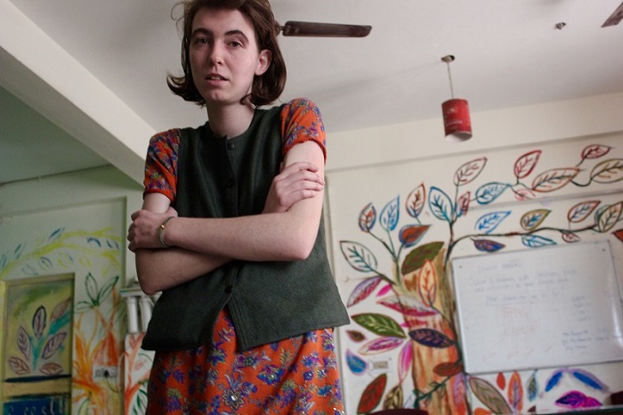 A self-conscious white woman with her arms crossed uncomfortably, wearing an Indian dress of bright orange-coloured suit with a green khadi jacket.