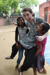 Image of a White woman with a small ponytail in "Indian dress code", wearing 'salwar-kameez' and being, to her surprise, lifted up by two grinning girl children.