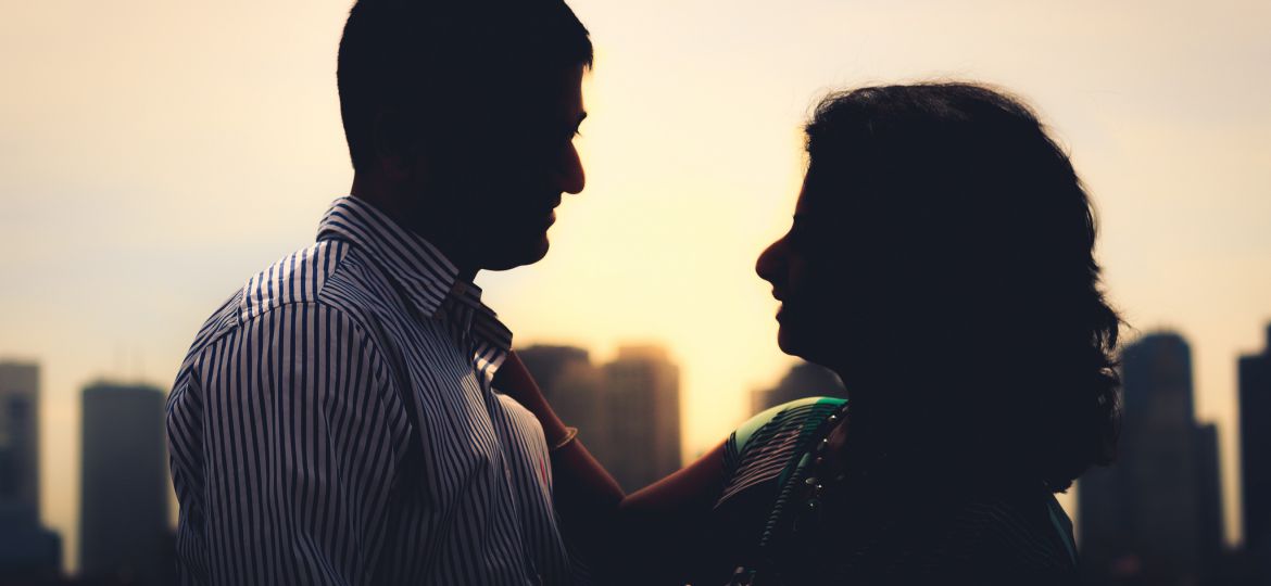 A man in white and blue striped shirt, and a woman in a green saree are standing on a terrace facing each other, with the woman's right hand on his left shoulder. Behind them are buildings, and a sun is setting down.