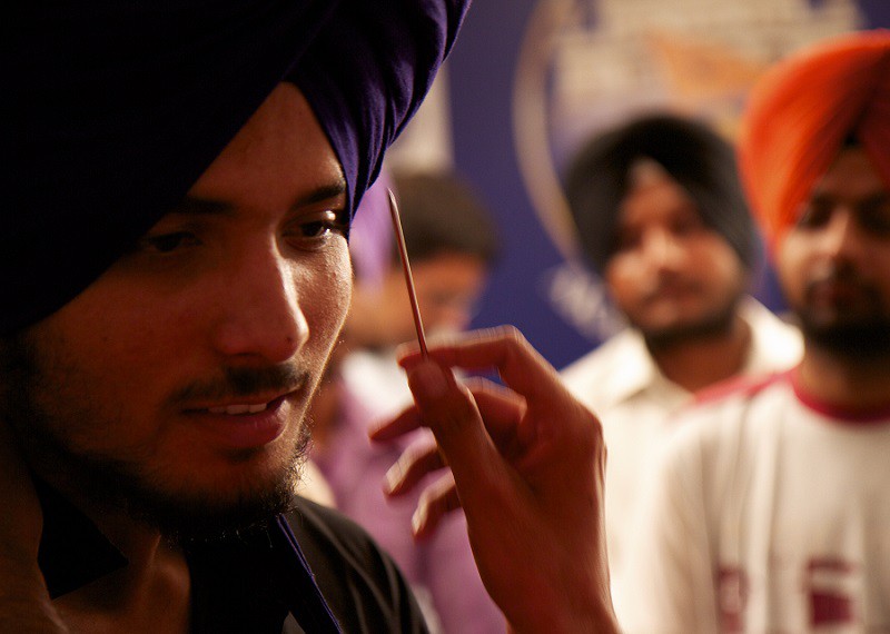 A close-up of the face of sikh man in a purple turban or dastar. Another person's hand pointing at his truban with a toothpick. Behind him are two other men in turban who are blurred out.