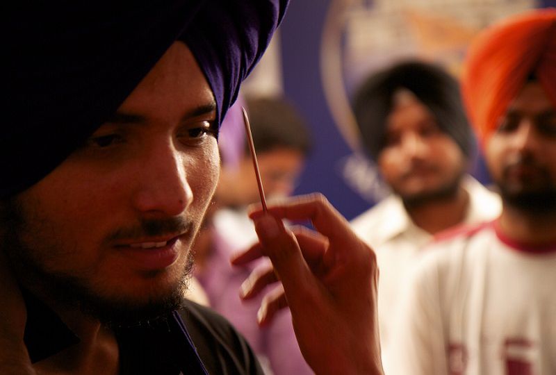 A close-up of the face of sikh man in a purple turban or dastar. Another person's hand pointing at his truban with a toothpick. Behind him are two other men in turban who are blurred out.