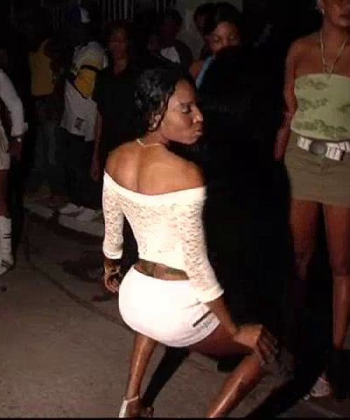 A black woman in a white shoulder top and white shorts dancing unfazed. She is the dancing pose where she is a little bent with her knees to the side and outward.