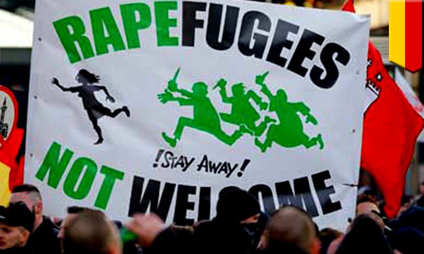 A photo of a protest with a focus on a placard that says in bold and all caps, "Rapefugess not welcome" with words "rape" and "not" in green, and the rest in black. In the middle is drawn a woman running away from a three men who are holding daggers in their hands, running towards her.