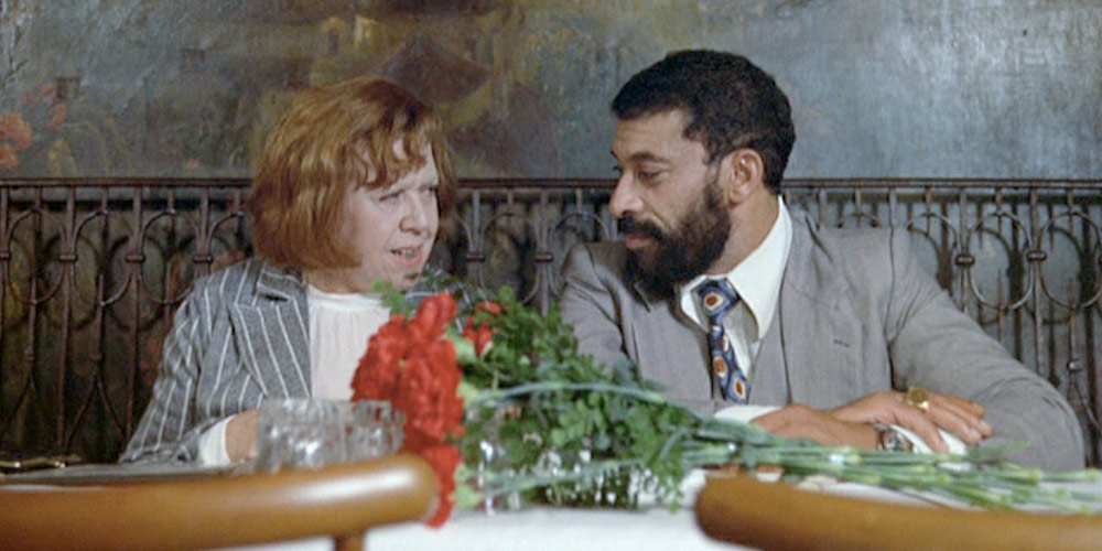 Still from a film. A man and a woman at a restaurant table, talking. A bunch of red flowers are kept on the table in front of them.