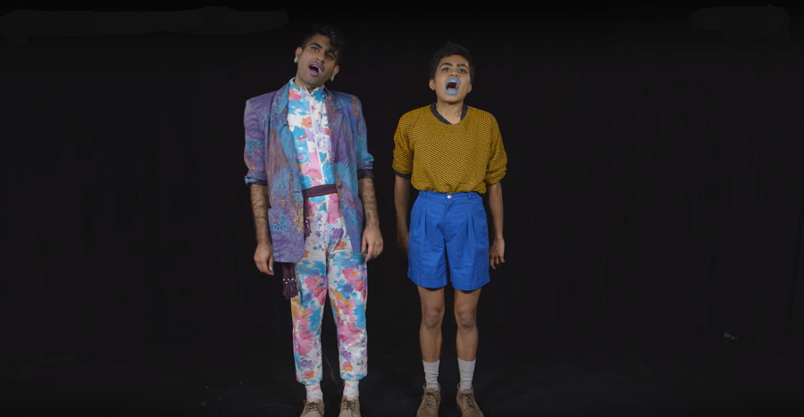 Photo of two brown, queer boys on a stage performing in front of a dark black background. One is wearing earrings, a blue overcoat, and a wite jumpsuit which has big baby pink and baby blue-colours flowers. Another is wearing bright blue lipstick, orange tee, and blue kneww-length shorts. They have their mouths wide open as if singing something.