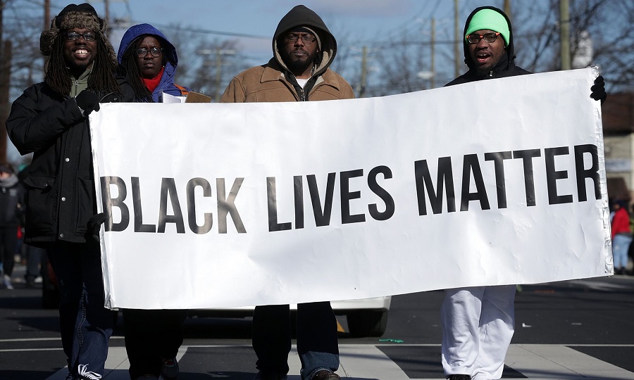 4 black men wearing jackets and caps carrying a big placard that says, "Black lives matter" written in black and bold on a white background.