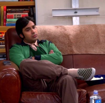 Still from American sitcom 'Big bang theory'. A brown Indian man in his late 20s, early 30s, sitting on a brown sofa with arms crossed, and one leg above the other, looking disconcerted.