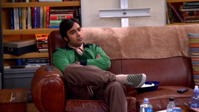 Still from American sitcom 'Big bang theory'. A brown Indian man in his late 20s, early 30s, sitting on a brown sofa with arms crossed, and one leg above the other, looking disconcerted.