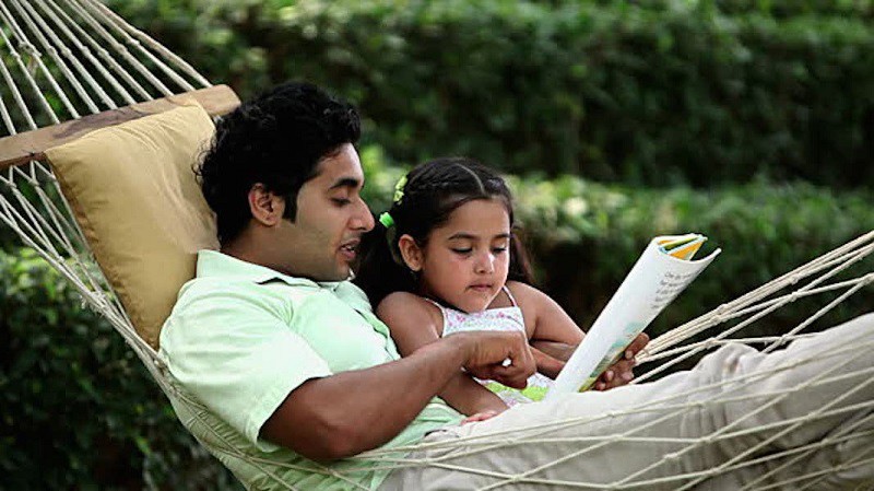 A father with his little daughter on a hammock, reading together from a book.