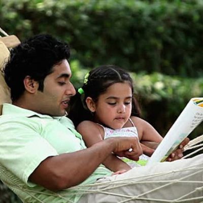 A father with his little daughter on a hammock, reading together from a book.