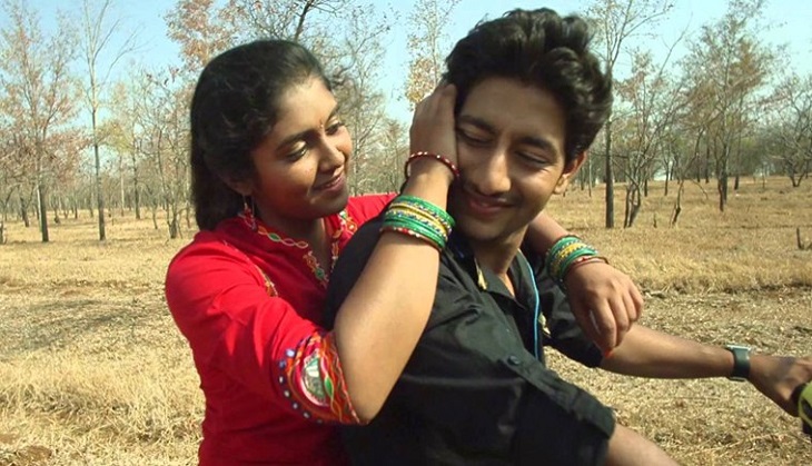 Still from film 'Sairat' (2016). A teenage girl sitting behind a teen boy on a two-wheeler. She has her arms on his shoulders, and is caressing his head with one hand. He is wearing a black shirt, and she is wearing a red ruit, green bangles, and a bindi on her forehead.