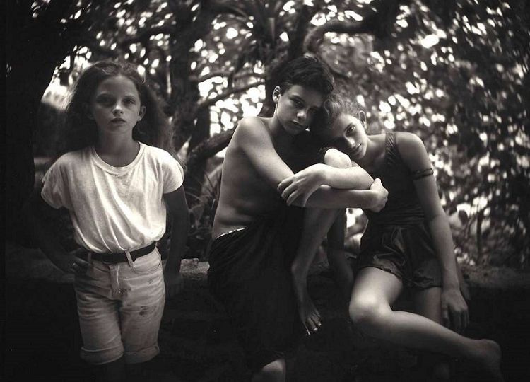 Black-and-white photo of three little children of about 5-7 years of age sitting on a branch of a tree. One girl is wearing knee-length shorts and tucked-in teeshirt; a boy wears a black knee-length shorts and and is naked above the waist; another girl resting her head on the boy's shoulder is wearing a thigh-length jumpsuit.