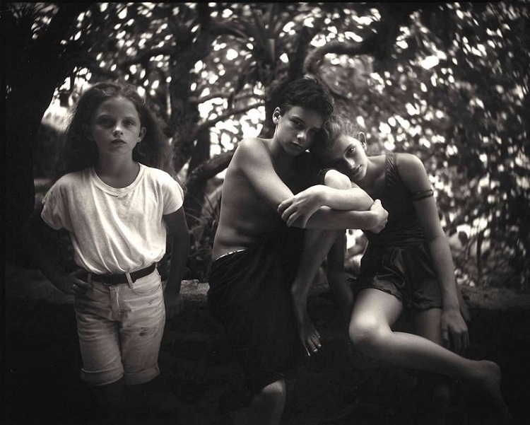 Black-and-white photo of three little children of about 5-7 years of age sitting on a branch of a tree. One girl is wearing knee-length shorts and tucked-in teeshirt; a boy wears a black knee-length shorts and and is naked above the waist; another girl resting her head on the boy's shoulder is wearing a thigh-length jumpsuit.