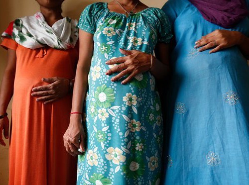 Photo of three Indian pregnant women from neck to knee with each resting their right hands on their protruding belly. One is wearin suit, and two are wearing night gown.