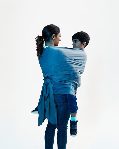 A mother wearing jeans and a tee carrying her school-age son in her arms. She has tied her dupatta around her and her son, and tied it behind her back so as to make a support to hold the child.