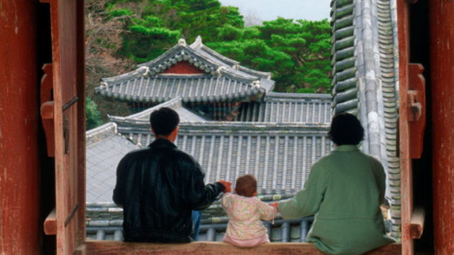 A family of a man, a child, and a woman sit on a window sill, facing mountains. They are holding the baby in between them - each parent holding his one hand.