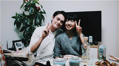 A chinese woman and man sitting in a living room of a house. The man has his left arm wrapped around her back. They are making the vicrtory symbol with their hands. The room has TV behind them, a dining table in front of them, and a room plant in a corner.