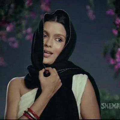 Still from a title song of Hindi film 'Satyam shivam sundaram'. A woman wearing light yellow-coloured saree with no blouse is clutching her dupatta that is covering her head. She is singing a song at night under a tree with small purple flowers.