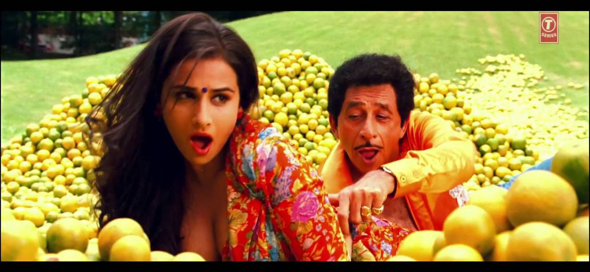 Still from a Hindi film 'The Dirty Picture' (2011). A brown woman has open black hair, is wearing a black bindi, and a bright-coloured, deep-neck blouse. A man behind her is touching her waist. There are piles of oranges in the garden around them. They both are singing a song.