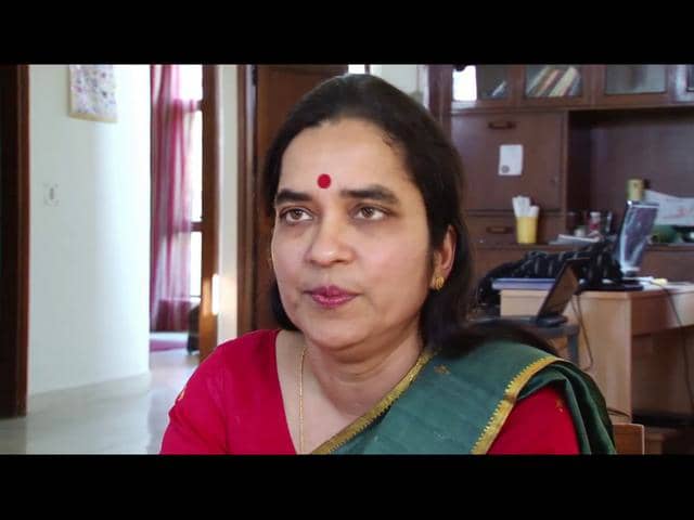 An Indian woman sitting in her speaking to a camera. She is wearing a green saree with red blouse, and a red bindi.