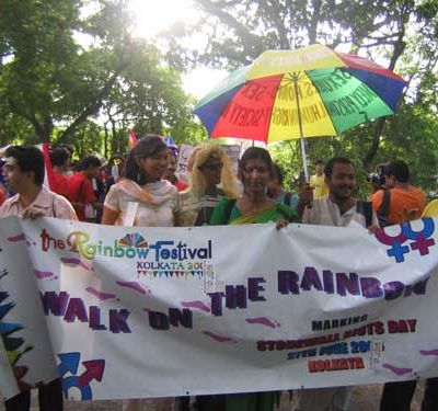 Photo from an Indian LGTB festival. Several people carrying together a big placard that reads, "The rainbow festival, Kolkata - Walk on the rainbow." One of them also carry a rainbow-coloured umbrella. The sun shines above.