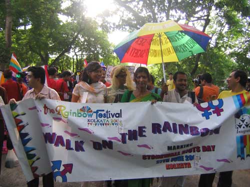 Photo from an Indian LGTB festival. Several people carrying together a big placard that reads, "The rainbow festival, Kolkata - Walk on the rainbow." One of them also carry a rainbow-coloured umbrella. The sun shines above.