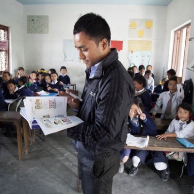 Photo of a young man as a school teacher in a classroom of little Nepalese children. He is thumbing through a picture book.