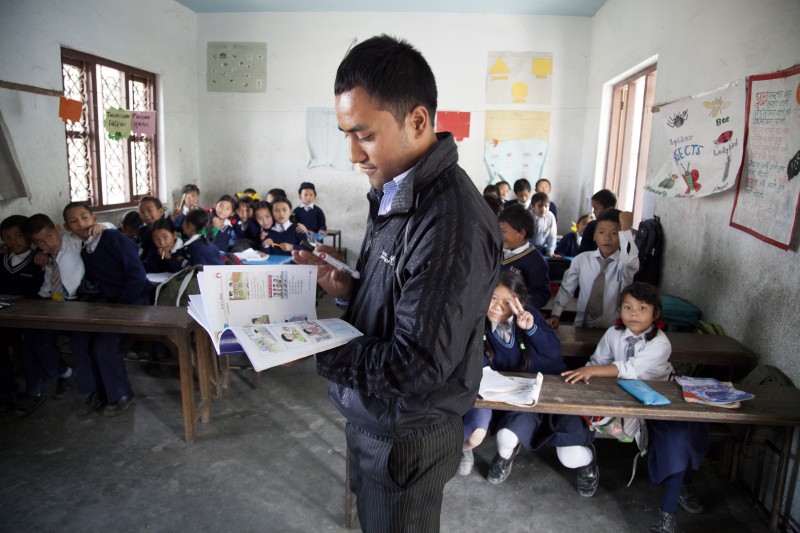 Photo of a young man as a school teacher in a classroom of little Nepalese children. He is thumbing through a picture book.