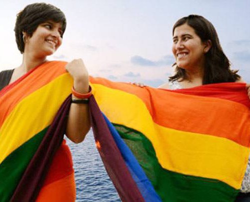 Two women stand side by side, one of them wears rainbow-coloured saree, and the one beside her drapes the pallu on her body