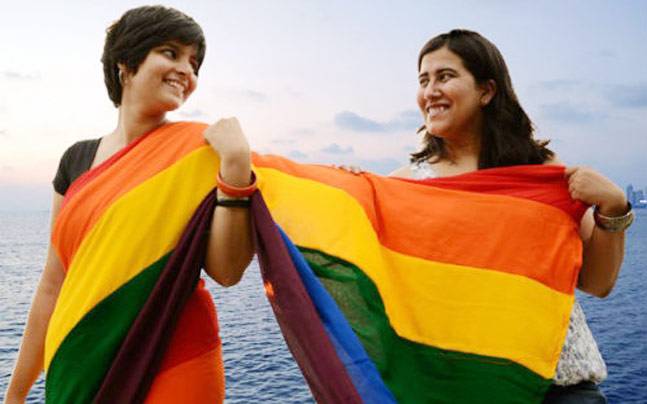 Two women stand side by side, one of them wears rainbow-coloured saree, and the one beside her drapes the pallu on her body