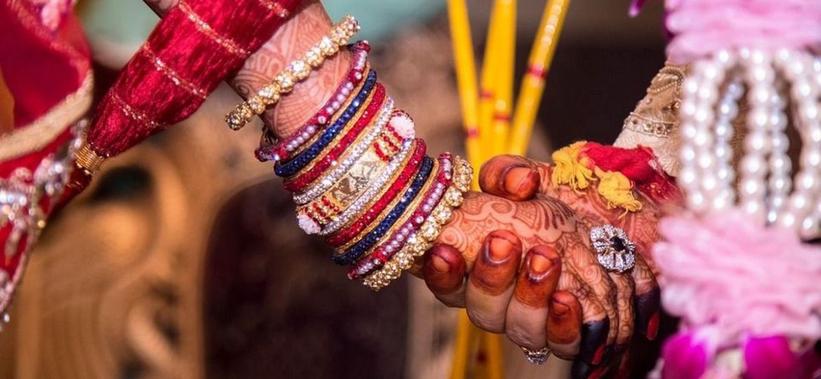 A zoomed in photograph of a couple holding hands during the marriage ceremony taking place. Both of them are wearing henna on their hands. The girl is wearing a set of bangles.