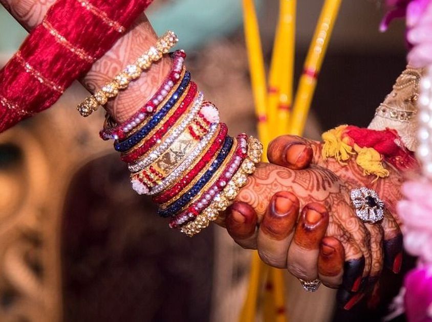 A zoomed in photograph of a couple holding hands during the marriage ceremony taking place. Both of them are wearing henna on their hands. The girl is wearing a set of bangles.