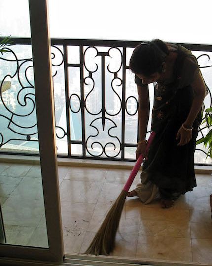 A photograph of a woman wearing a green saree cleaning the balcony with a broom.