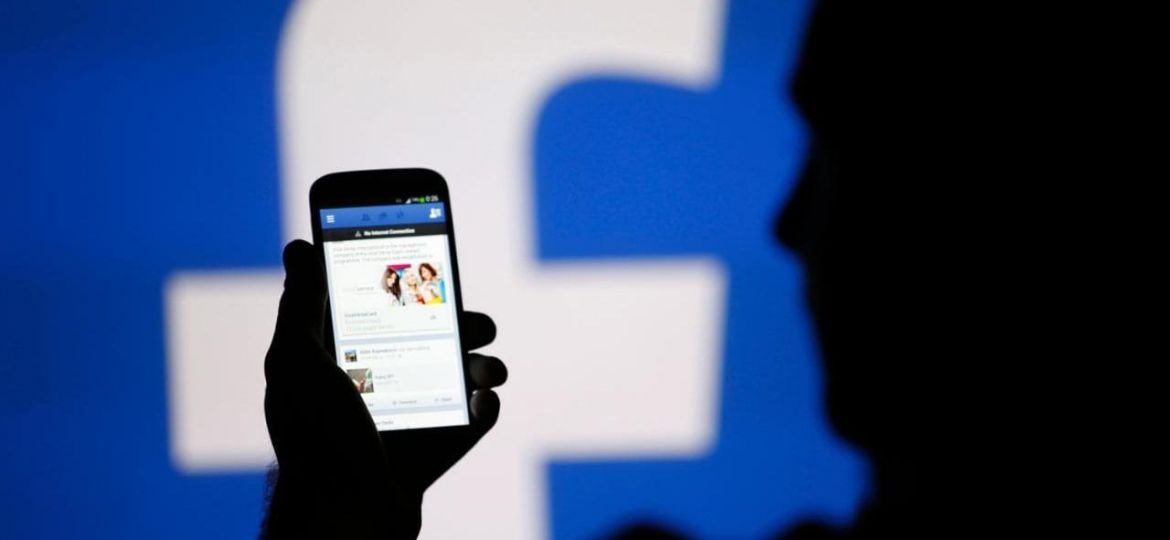 The background of the photo has the 'f' symbol of faacebook. The image features a a person (silhouette) looking at their phone, which shows the news feed page of Facebook app.