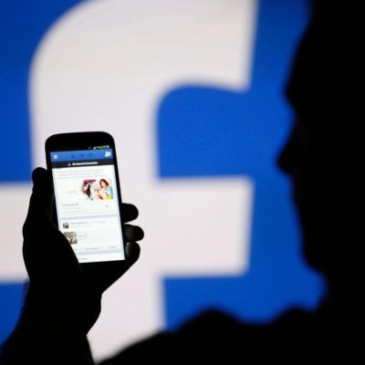 The background of the photo has the 'f' symbol of faacebook. The image features a a person (silhouette) looking at their phone, which shows the news feed page of Facebook app.