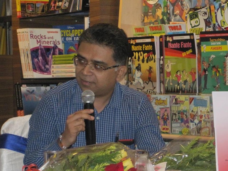 A photo of Pawan Dhall wearing a blue shirt, speaking on the mike with book shelves in the background