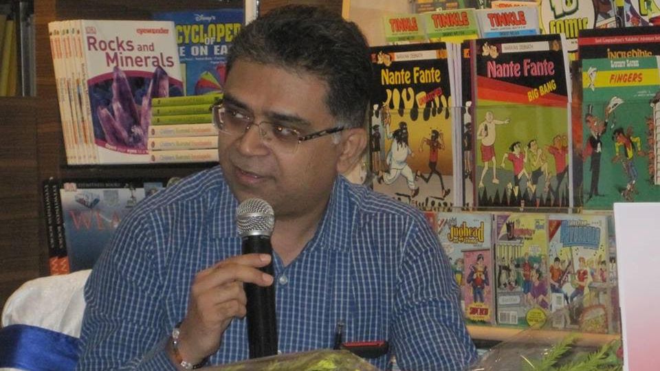 A photo of Pawan Dhall wearing a blue shirt, speaking on the mike with book shelves in the background