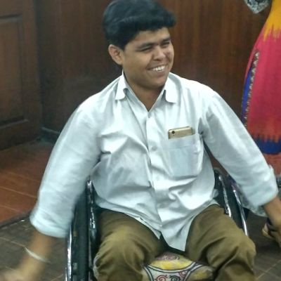 Kiran sitting on a wheelchair, smiling. He is wearing brown pants and a white shirt, and has his cellphone peeking out of his top shirt pocket.