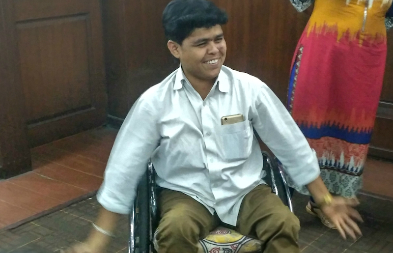 Kiran sitting on a wheelchair, smiling. He is wearing brown pants and a white shirt, and has his cellphone peeking out of his top shirt pocket.