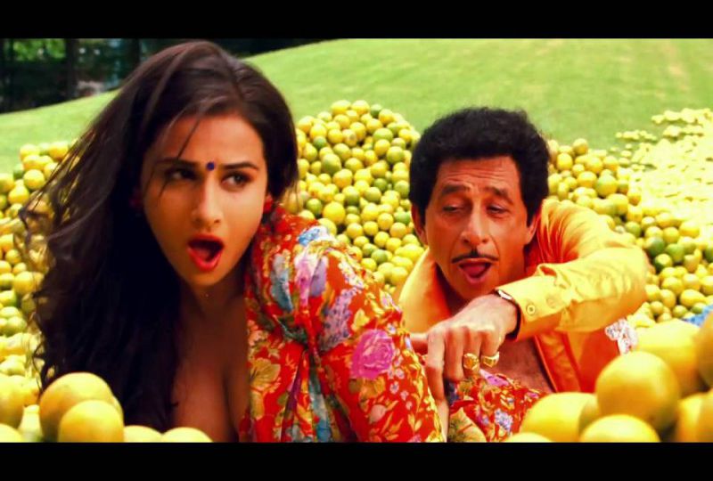 Still from a Hindi film 'The Dirty Picture' (2011). A brown woman has open black hair, is wearing a black bindi, and a bright-coloured, deep-neck blouse. A man behind her is touching her waist. There are piles of oranges in the garden around them. They both are singing a song.
