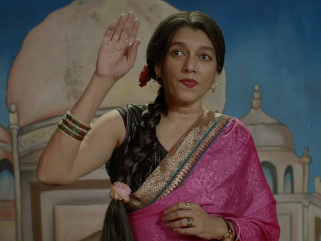 Still from a Hindi film 'Lipstick under my Burkha'. The character of Ratna Pathak Shah is waving so as to get someone's attention. She is wearing a rose in her hair, a pink saree with golden border, black blouse, and has her hair tied in a plait that has been pulled to the front. She is wearing coloured bangles, and earrings.