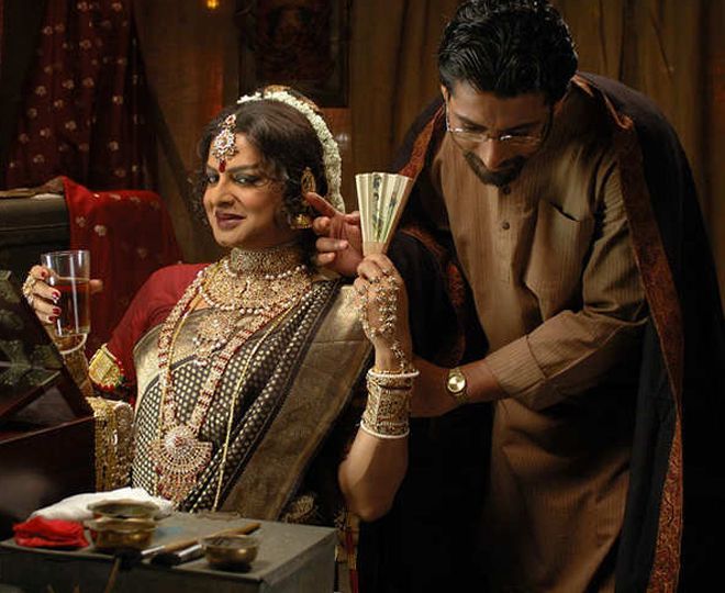 A still from a 2010 film 'Just Another Love Story'. A middle-aged man dressed in brown kurta-pajama is bowing as if in courtsey to a woman who is wearing heavy jewellery. She is holding a glass of wine or water in one hand, and chinese fan in another. She is wearing a big red bindi, dark mehroon lipstick, and kajal.