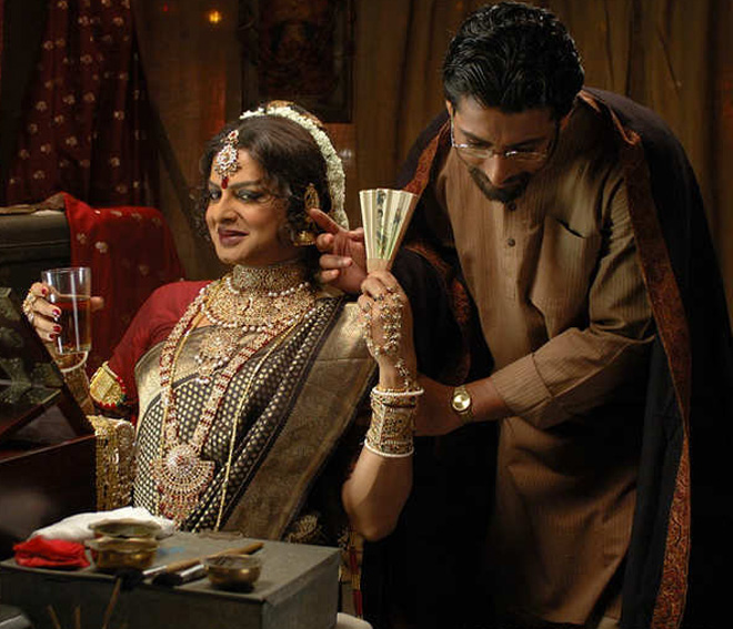 A still from a 2010 film 'Just Another Love Story'. A middle-aged man dressed in brown kurta-pajama is bowing as if in courtsey to a woman who is wearing heavy jewellery. She is holding a glass of wine or water in one hand, and chinese fan in another. She is wearing a big red bindi, dark mehroon lipstick, and kajal.