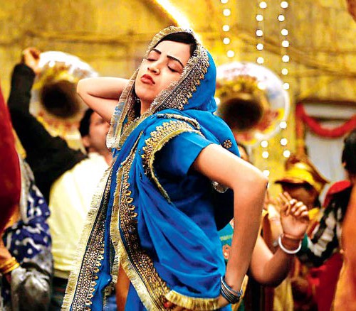 Still from a Hindi film 'Lipstick under my Burkha'. A young woman wearing blue-coloured lehnga is dancing merrily in an Indian wedding. She has one hand on her hip, another behind her head, and is tilting upwards with her eyes closed, and lips pout.